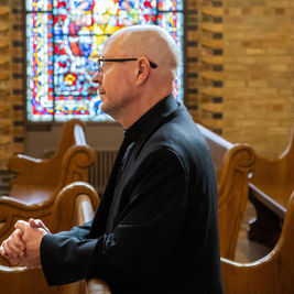 Bald man in black clerical attire sits in a church pew beside a stained-glass window, hands clasped in quiet reflection.