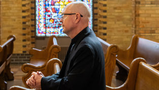 Bald man in black clerical attire sits in a church pew beside a stained-glass window, hands clasped in quiet reflection.