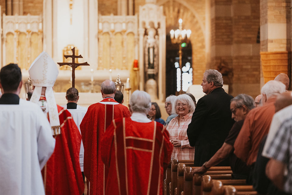 Congregation stands in a church service as clergy in red robes face the altar; an elderly woman smiles near the pews.