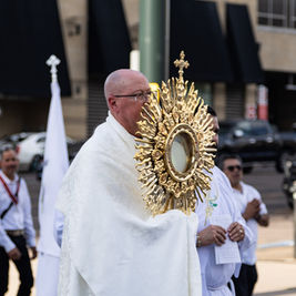 Priest in white robes carries a gold monstrance in a street procession, with flags and other participants behind him.