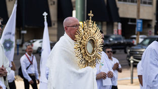 Priest in white robes carries a gold monstrance in a street procession, with flags and other participants behind him.