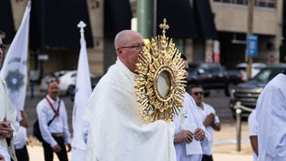 Priest in white robes carries a gold monstrance in a street procession, with flags and other participants behind him.