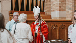 Smiling bishop in red vestments greets churchgoers in a church, holding a staff near wooden pews and organ pipes.