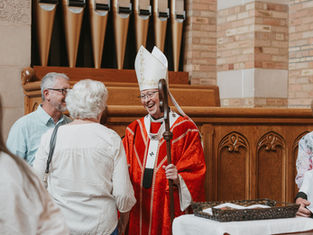 Smiling bishop in red vestments greets churchgoers in a church, holding a staff near wooden pews and organ pipes.