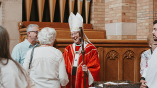 Smiling bishop in red vestments greets churchgoers in a church, holding a staff near wooden pews and organ pipes.