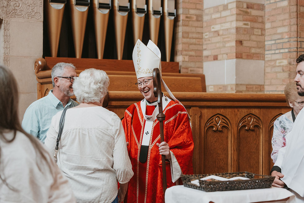 Smiling bishop in red vestments greets churchgoers in a church, holding a staff near wooden pews and organ pipes.