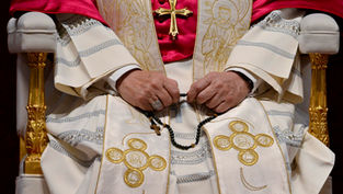 Pope seated in ornate white and red vestments, hands holding a rosary, with a gold cross and embroidered religious motifs
