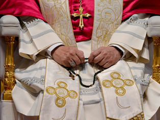 Pope seated in ornate white and red vestments, hands holding a rosary, with a gold cross and embroidered religious motifs