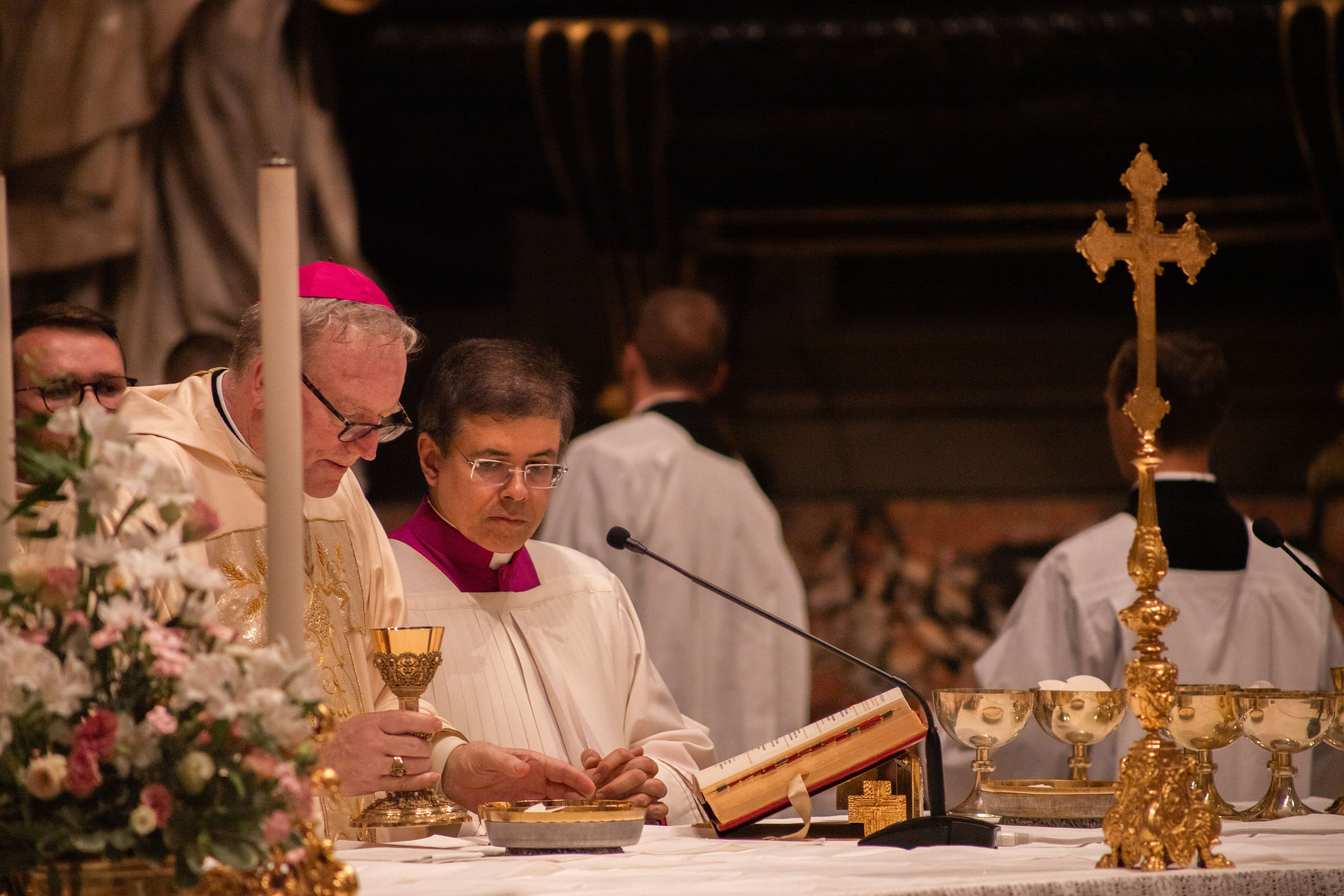 Catholic bishops at a candlelit altar during Mass, reading from a book beside a golden cross and chalices.