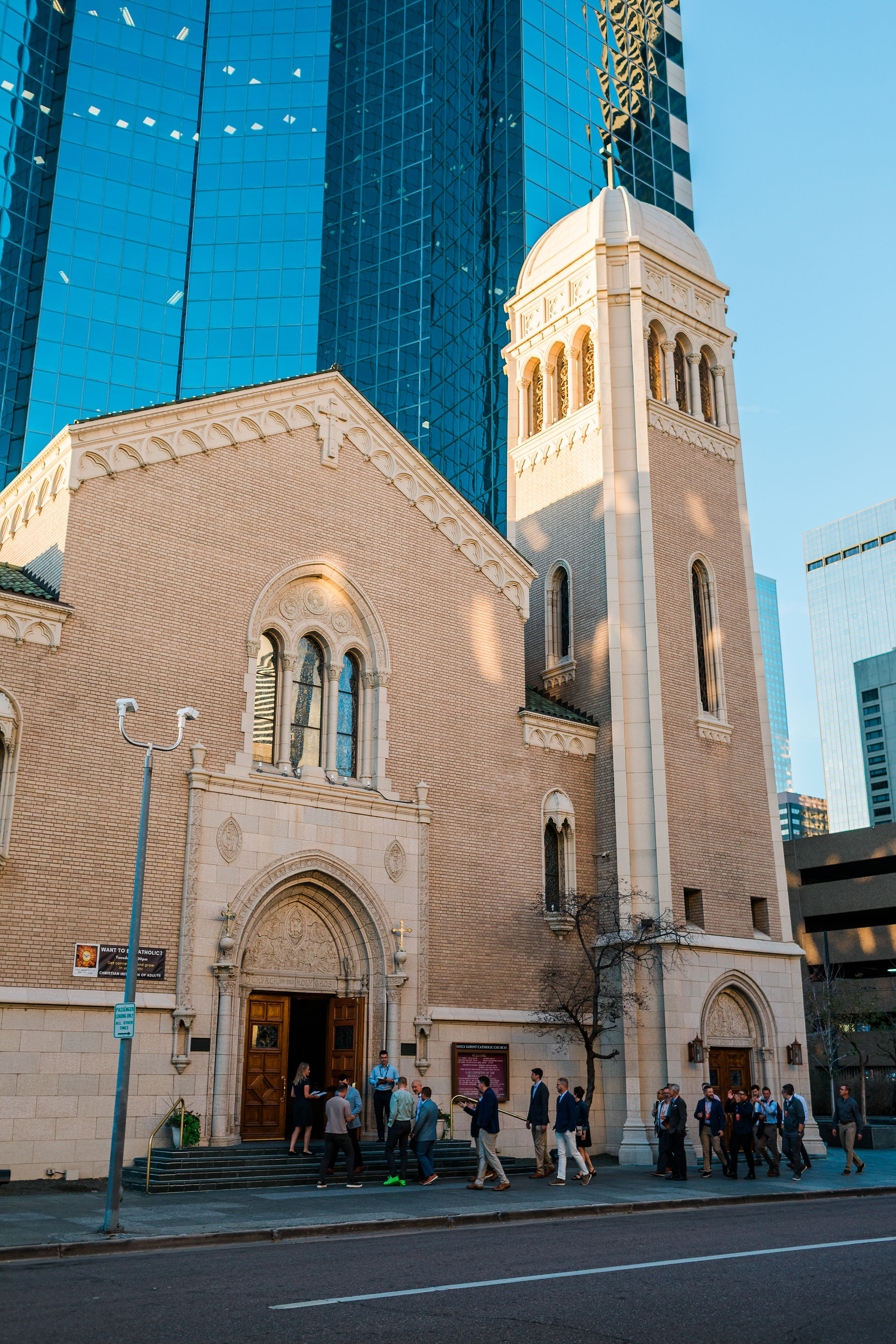 People gather outside a beige church with a tall tower, set against blue glass skyscrapers in warm late-afternoon light.