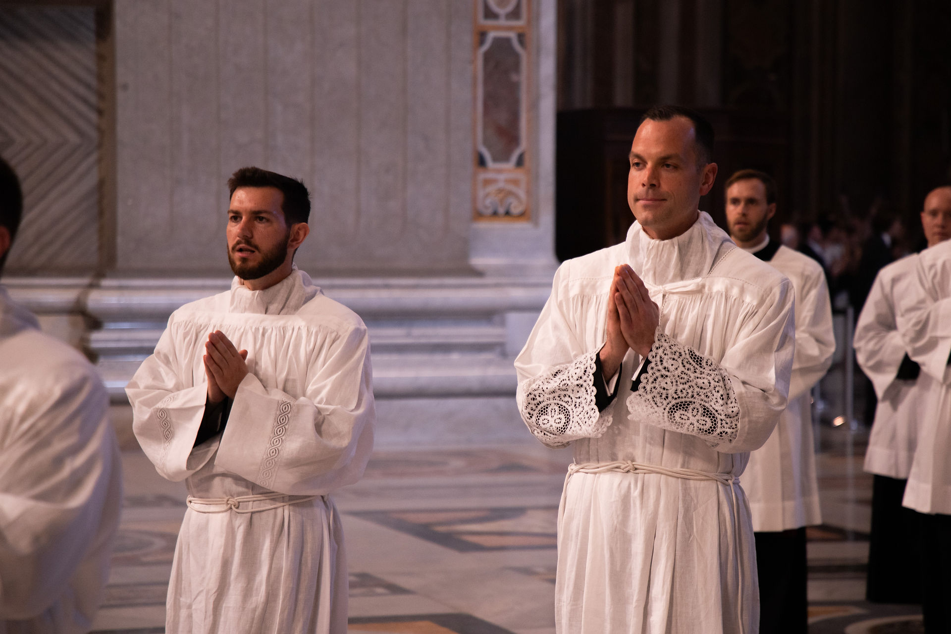 Men in white robes stand with hands clasped in prayer inside a cathedral during a solemn ceremony.