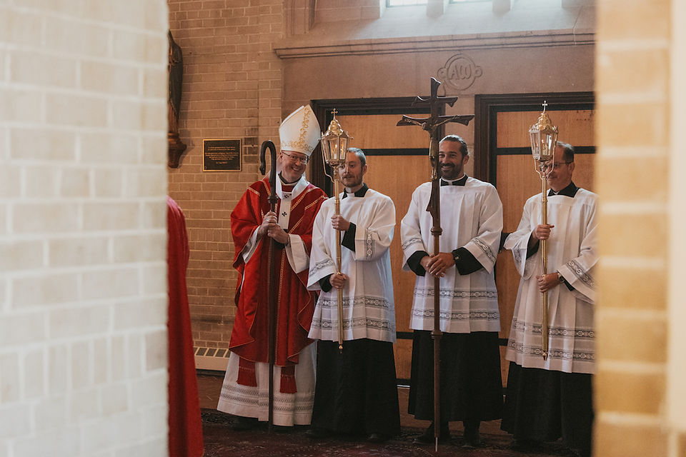 Clergy in red and white robes stand in a church entrance holding candles and a crucifix, smiling during a solemn procession.
