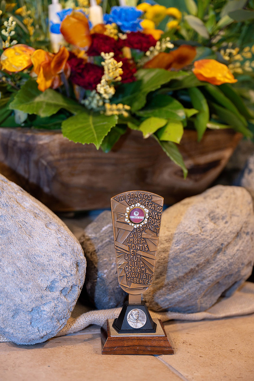 Bronze reliquary with Latin inscription on a wood base, set among rocks before colorful flowers and greenery.