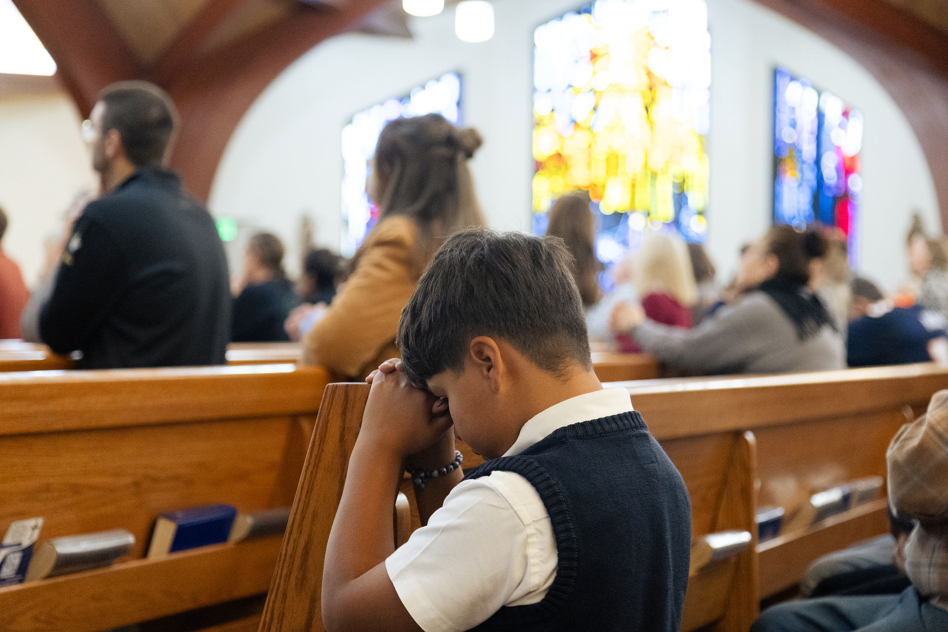 Boy kneels praying in a church pew, hands clasped, with blurred worshippers and bright stained glass in the background.