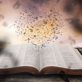 Open Bible on a wooden table with black letters rising into a cloudy sunset sky, suggesting inspiration and reverence
