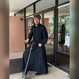 Priest in black cassock and bike helmet stands on a scooter outside a glass building entrance, with a poster by the door.
