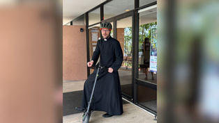 Priest in black cassock and bike helmet stands on a scooter outside a glass building entrance, with a poster by the door.