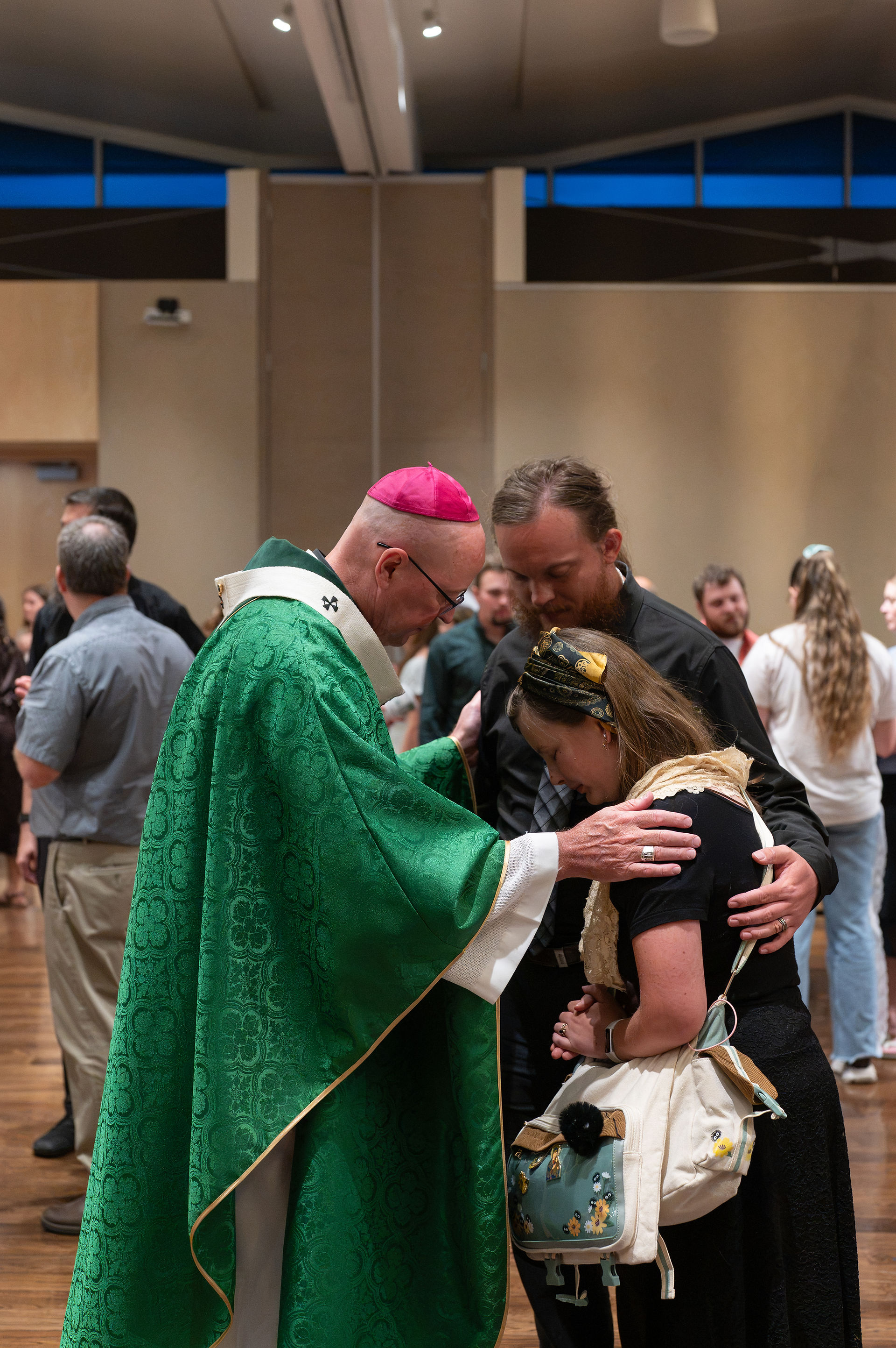 Priest in green vestments blesses a woman and man in a hall, all heads bowed in a quiet, solemn moment