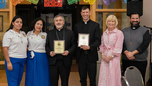 Six adults pose indoors under colorful papel picado, two priests holding plaques; all smiling at a church ceremony.