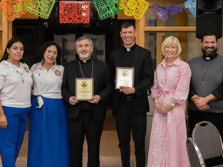 Six adults pose indoors under colorful papel picado, two priests holding plaques; all smiling at a church ceremony.