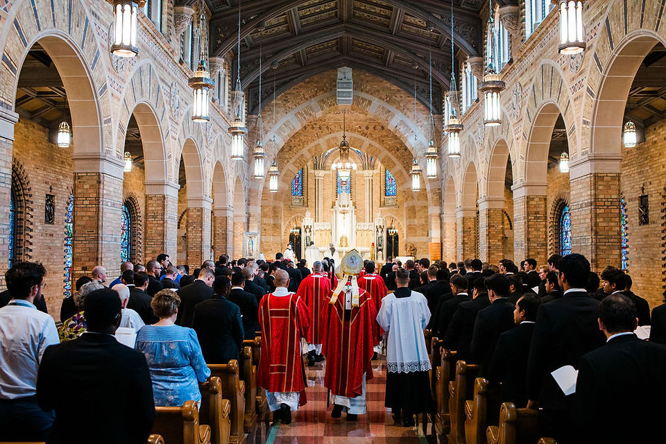 Congregation and priests in red vestments process down a grand brick church aisle toward the altar during a solemn service.