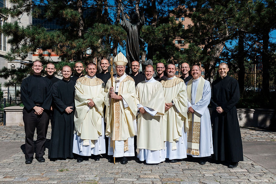 Group of Catholic priests and clergy pose outdoors in black and white vestments beneath trees and a statue.