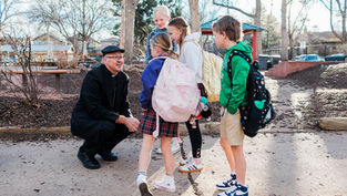 Priest kneels talking with four backpacked schoolchildren in a sunny park playground, creating a friendly, calm scene.