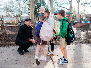 Priest kneels talking with four backpacked schoolchildren in a sunny park playground, creating a friendly, calm scene.
