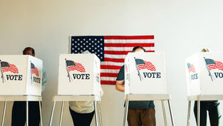 Voters stand behind white booths marked VOTE in front of a U.S. flag at a polling place.