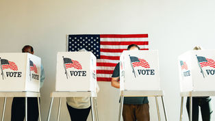 Voters stand behind white booths marked VOTE in front of a U.S. flag at a polling place.
