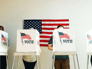 Voters stand behind white booths marked VOTE in front of a U.S. flag at a polling place.