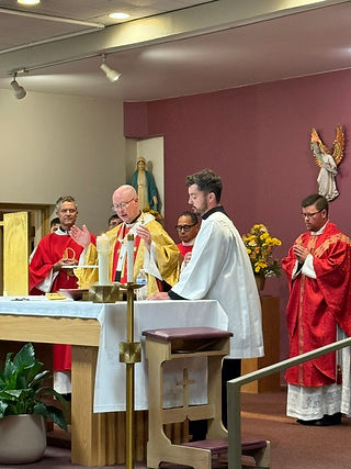 Priests in red and white vestments celebrate Mass at an altar in a chapel, with candles, flowers, and statues in the background.