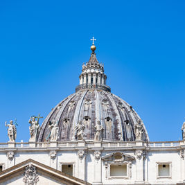 St. Peter’s Basilica dome and statues against a clear blue sky, with a clock on the right and no people visible.