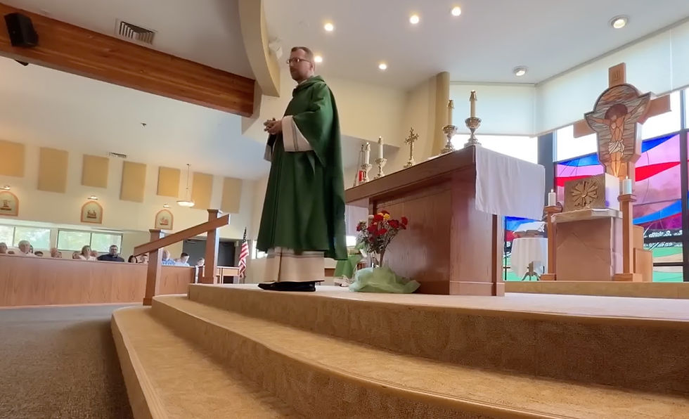 Priest in green vestments stands on altar steps in a bright church, with candles, stained glass, and seated parishioners behind.