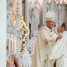 Bishop in cream vestments greets a new deacon inside an ornate white Gothic church, smiling during a ceremony.