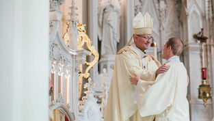 Bishop in cream vestments greets a new deacon inside an ornate white Gothic church, smiling during a ceremony.