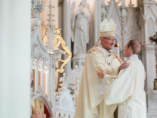 Bishop in cream vestments greets a new deacon inside an ornate white Gothic church, smiling during a ceremony.