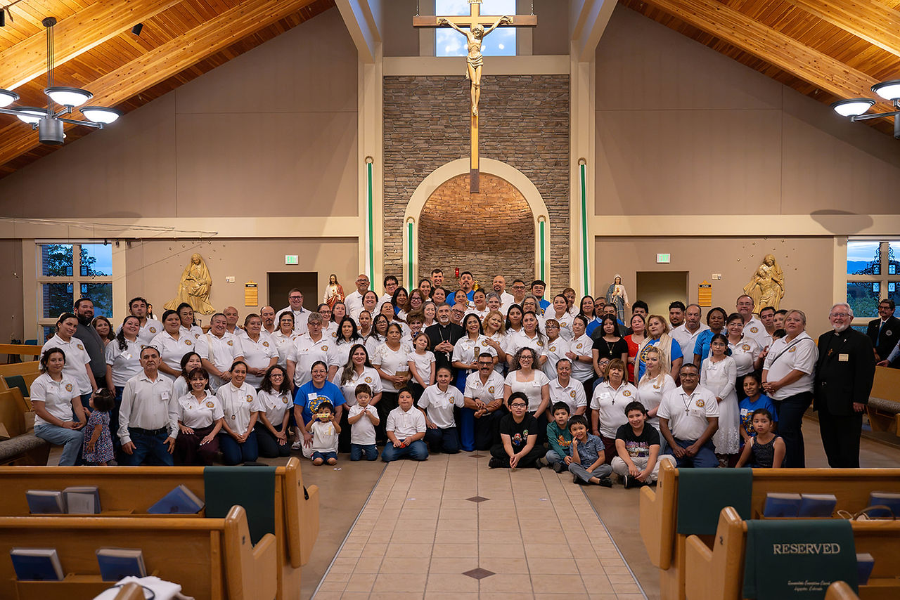 Large group posing inside a church beneath a crucifix, many in white shirts, with pews and statues in warm wood and stone interior