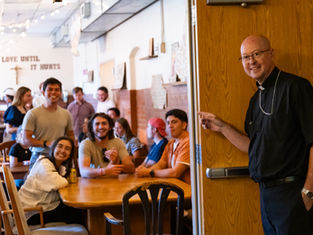 Smiling priest points into a lively church hall where young adults sit at wooden tables under the words LOVE UNTIL IT HURTS.