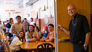 Smiling priest points into a lively church hall where young adults sit at wooden tables under the words LOVE UNTIL IT HURTS.