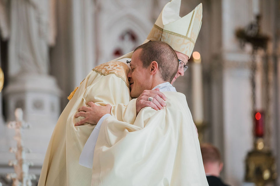 Bishop in cream vestments hugs a man in white robes inside a church, a warm, emotional moment during a service.