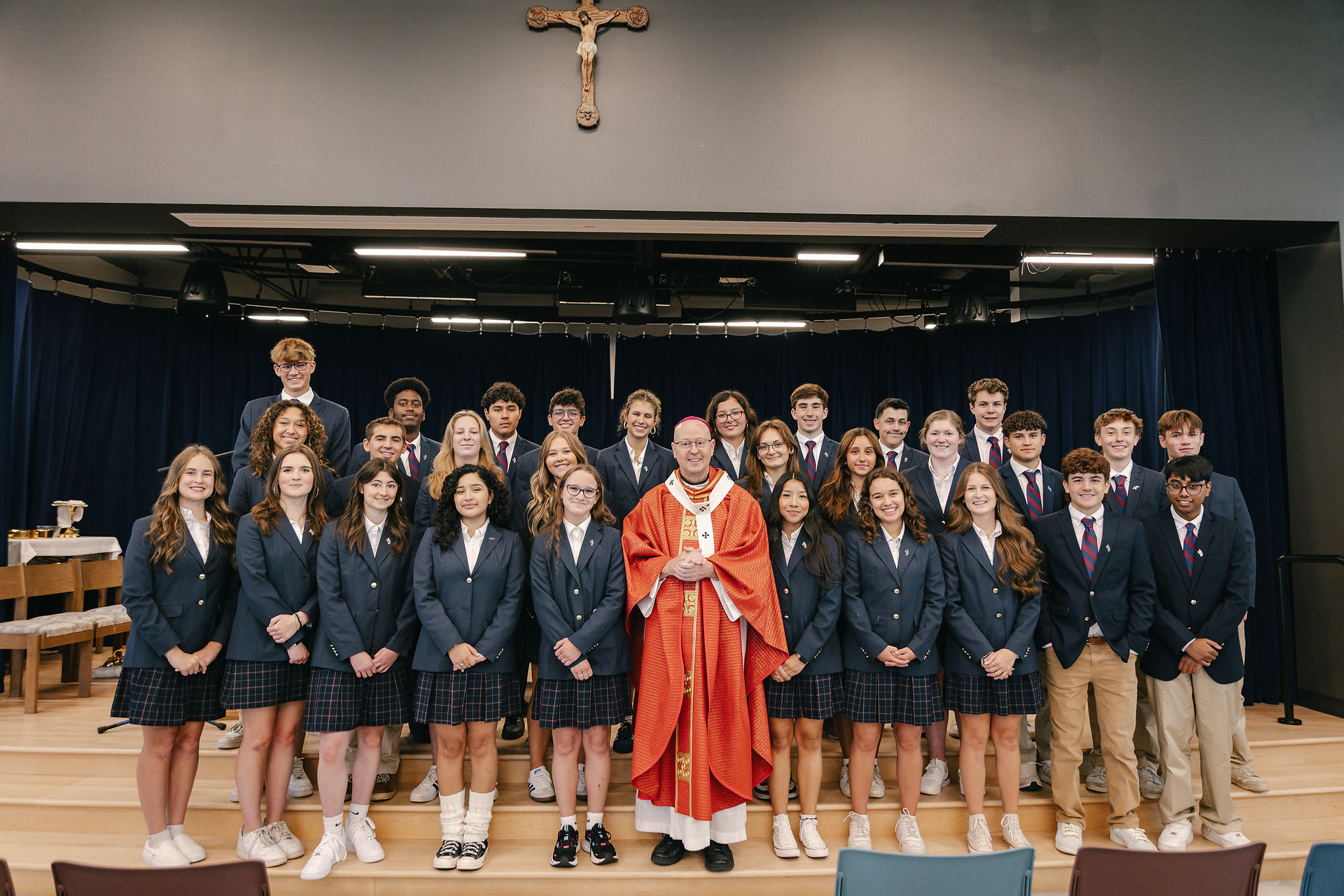 Students in school uniforms pose with a bishop in red vestments on a chapel stage beneath a crucifix, all smiling.