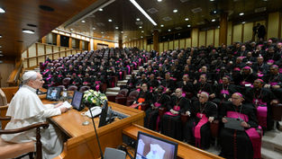 Pope speaks at a podium to a large seated group of bishops in black robes with pink sashes inside a wood-paneled auditorium.