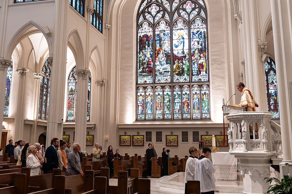 Priest in white and gold robe speaks at pulpit as congregation stands in a bright Gothic church with stained glass windows.