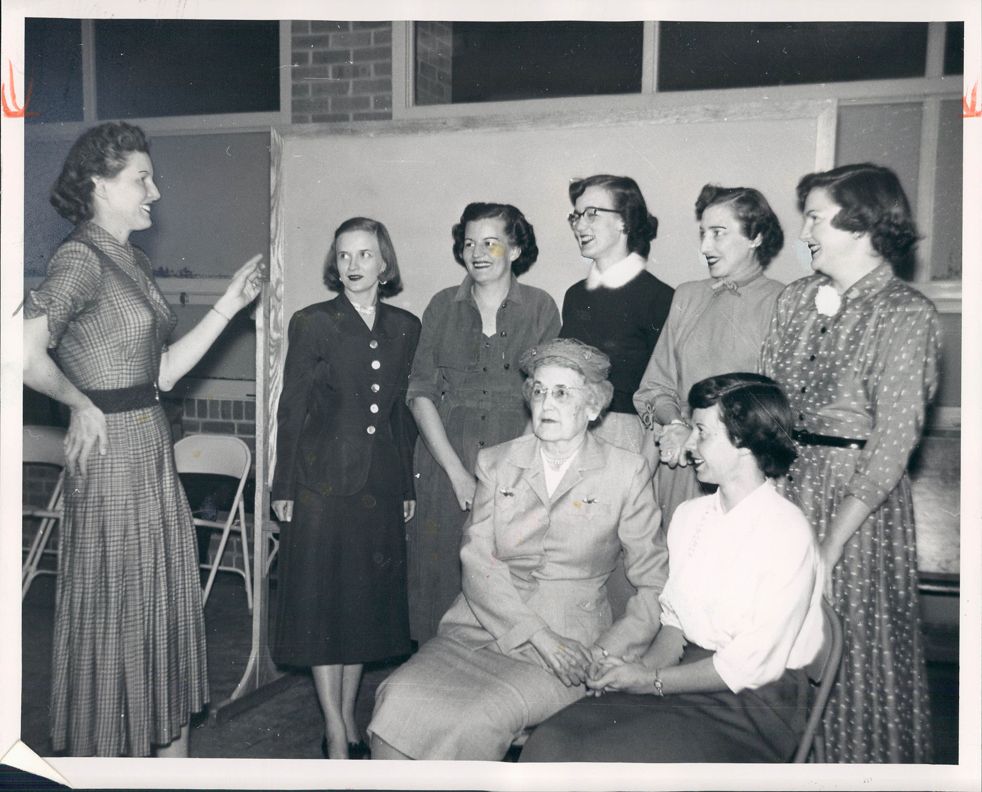 Black-and-white photo of seven women gathered indoors; one gestures as the others smile and listen in a classroom-like room.
