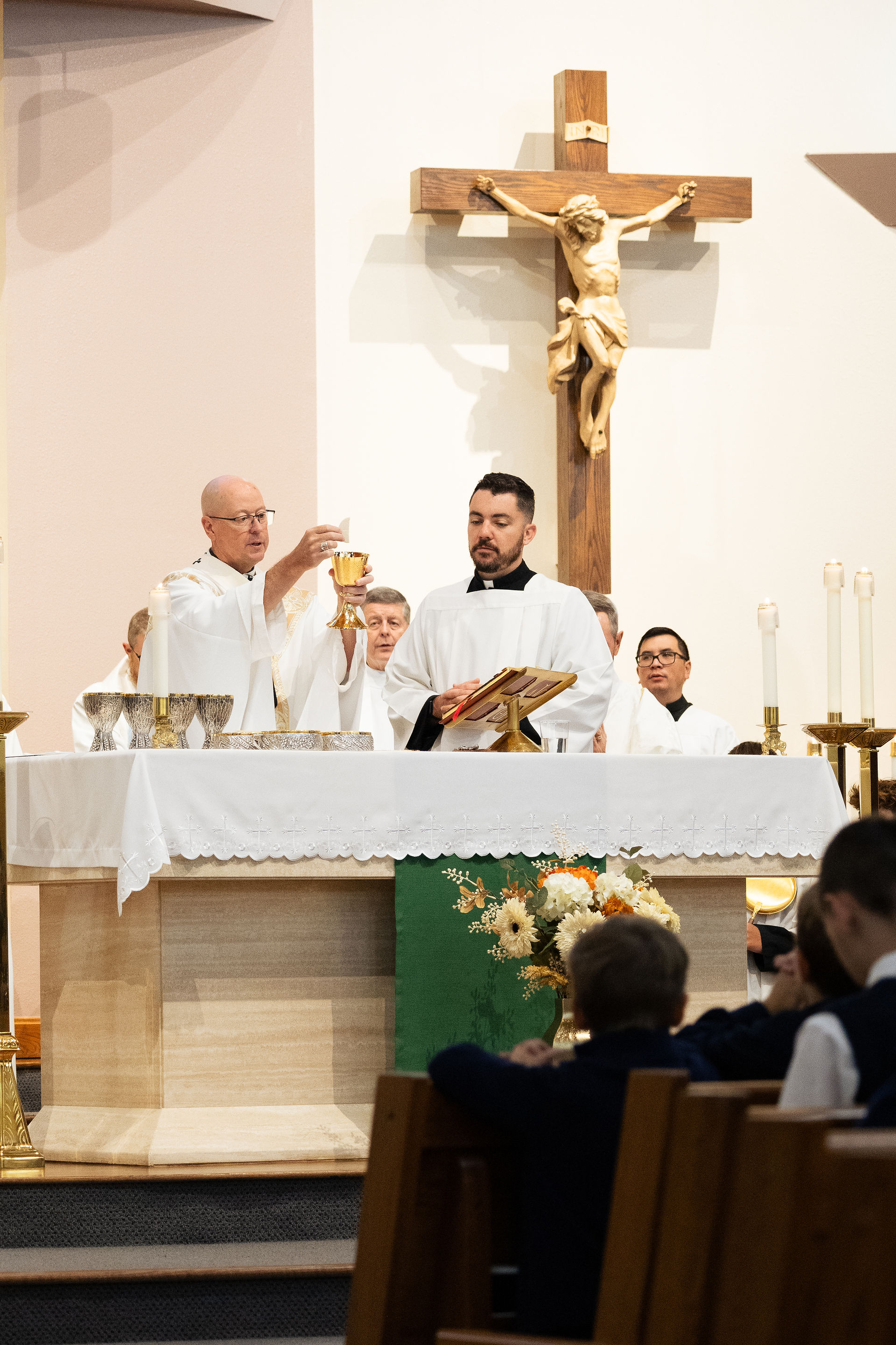 Priests in white vestments celebrate Mass at an altar beneath a large crucifix, with candles, chalice, and seated worshippers.