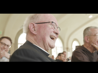 Elderly man in glasses laughing with others in an arched, bright room. Warm atmosphere, joy visible on their faces.