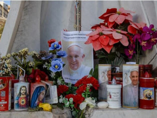 Flowers, candles, and photos of a religious figure create a vibrant memorial on a stone surface, conveying a solemn and respectful mood.