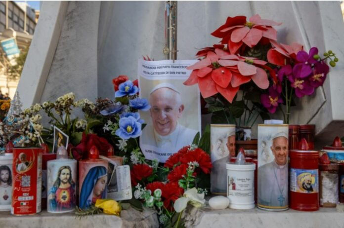 Flowers, candles, and photos of a religious figure create a vibrant memorial on a stone surface, conveying a solemn and respectful mood.