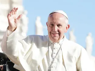A man in white religious attire waves, smiling outdoors against a clear blue sky. Statues in the blurred background.